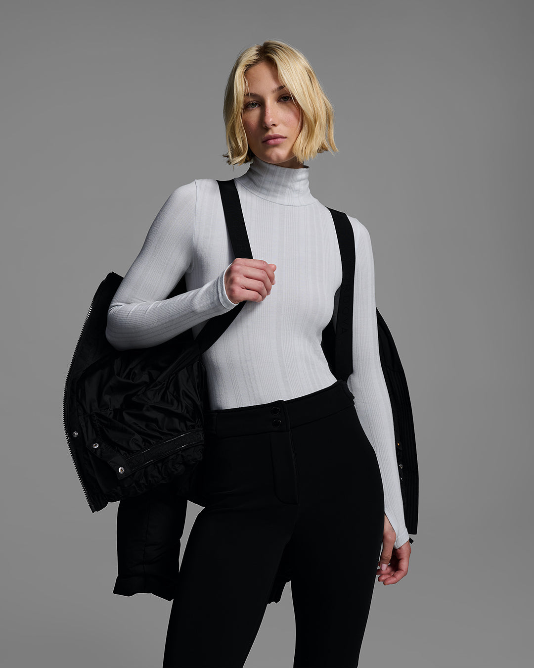 Knee up detail shot of a female model wearing a classic down filled ski jacket open, worn like a backpack in black with a matching ski pant and gray base layer. #color_onyx