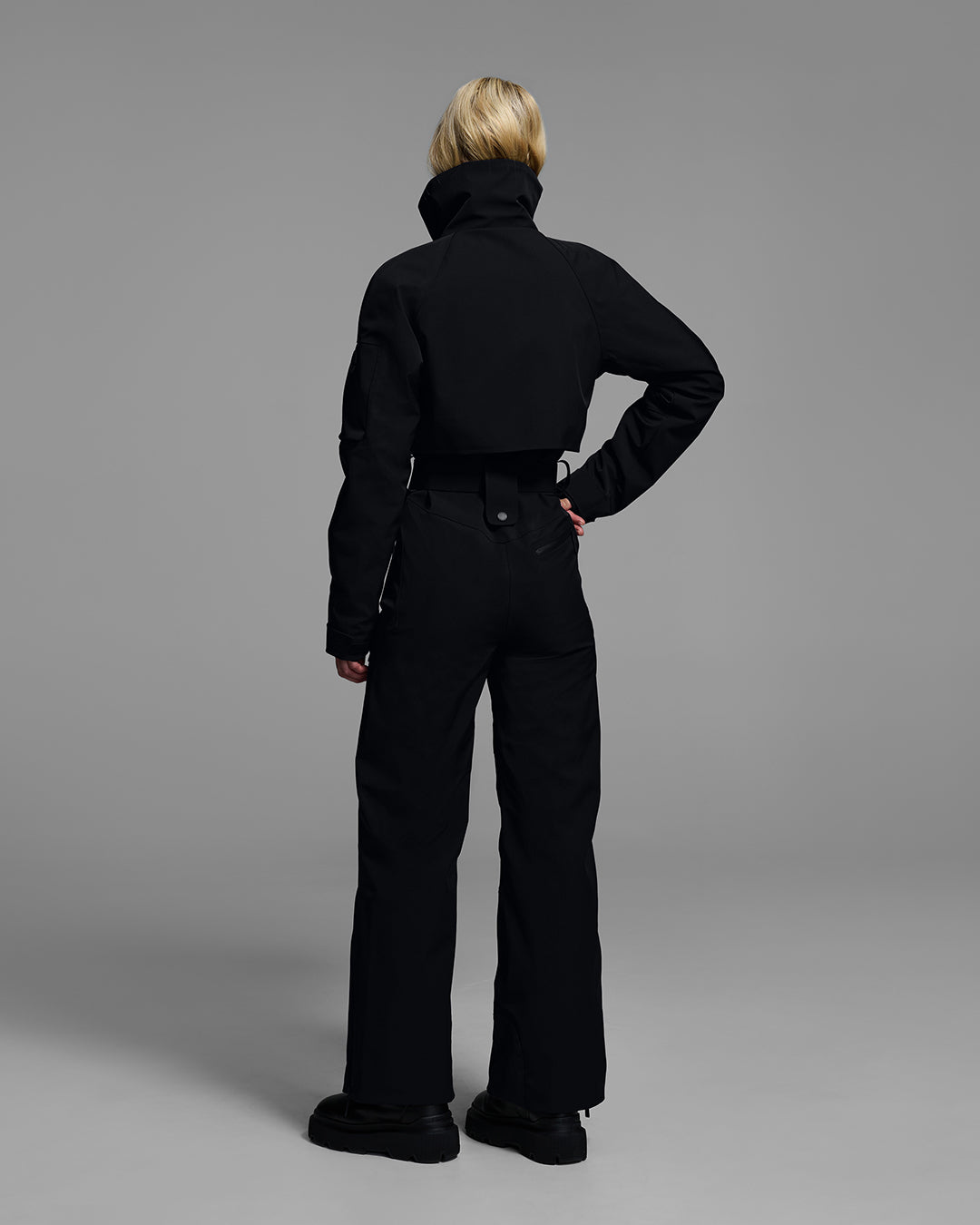 Back view of a woman wearing a black color waterproof technical fabric ski suit with a belted waist. #color_onyx