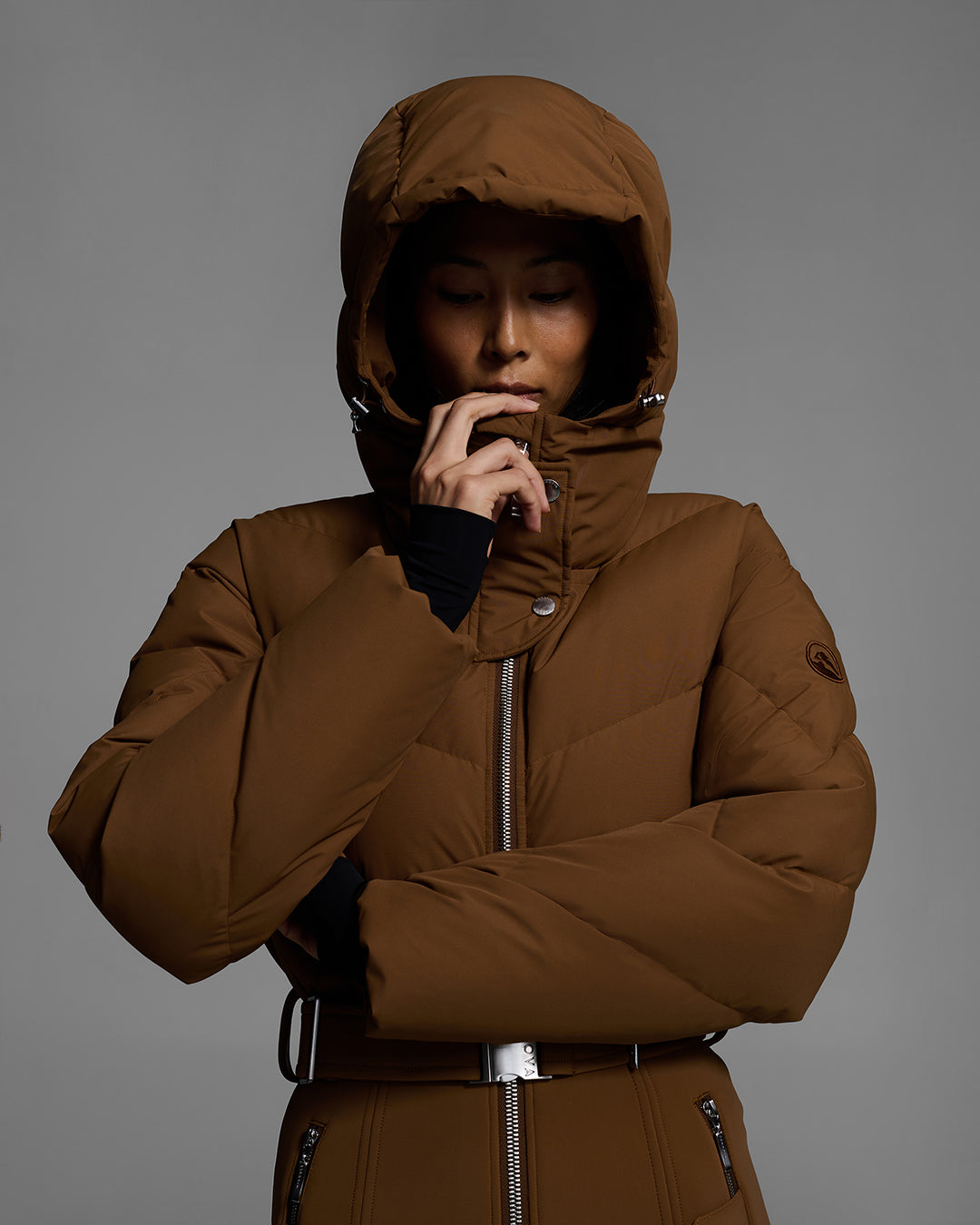 Waist up view of a female model wearing a belted sepia brown color ski suit with down filled top and 4-way stretch bottom. Her hood is on. #color_sepia