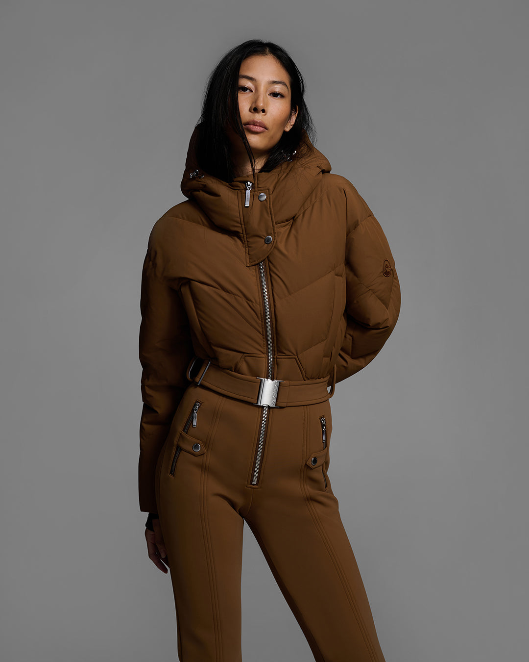 Knees up view of a female model wearing a belted sepia brown color ski suit with down filled top and 4-way stretch bottom. #color_sepia