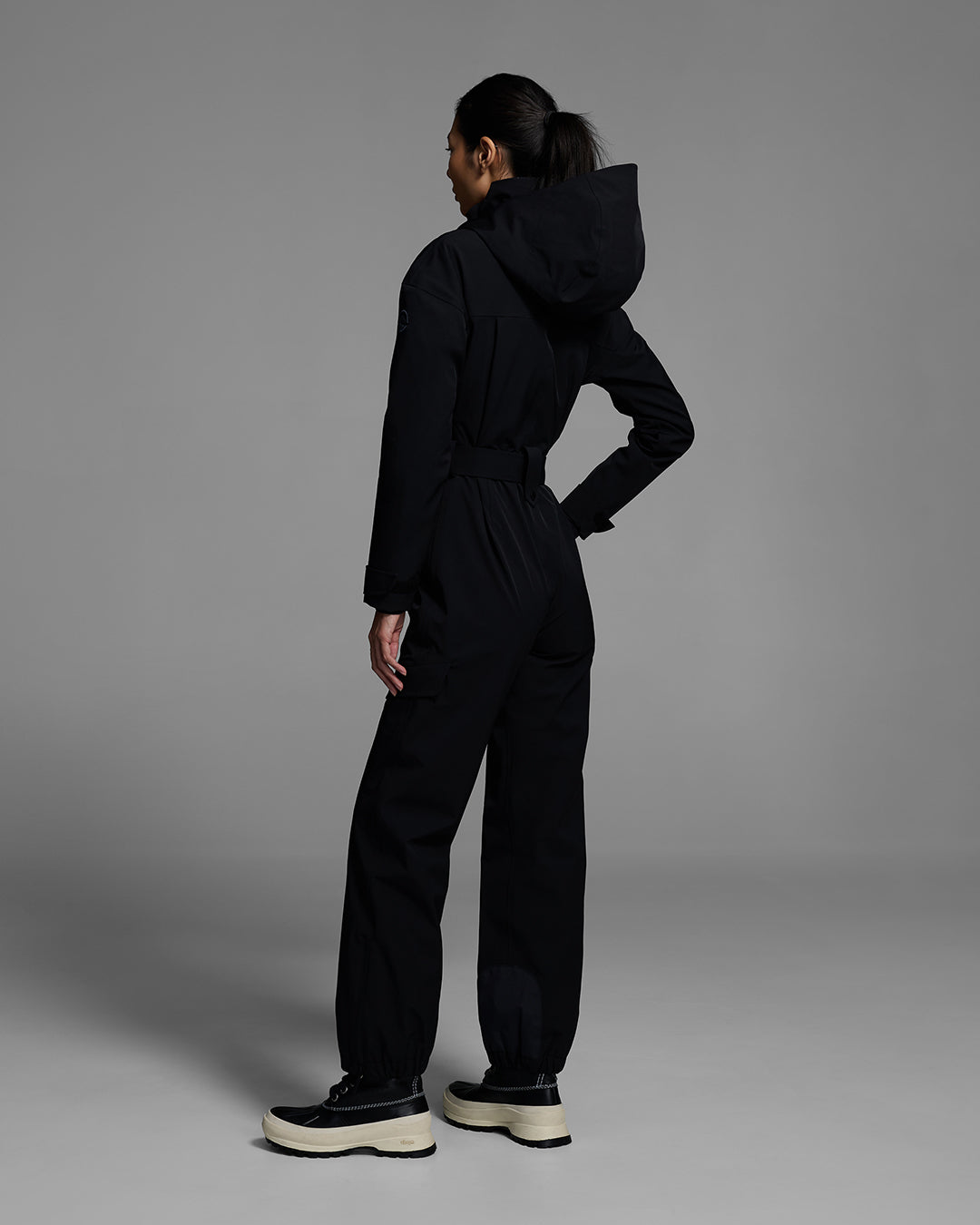 Back view of a female model wearing a belted onyx black color ski suit made of technical waterproof fabric and zippers. #color_onyx