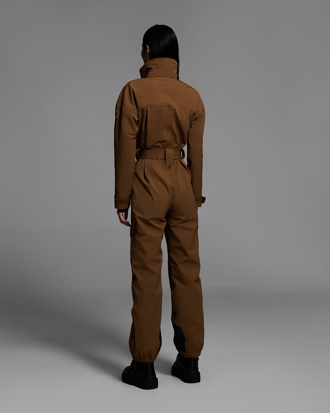 Back view of a female model wearing a belted sepia brown color ski suit made of technical waterproof fabric and zippers. #color_sepia