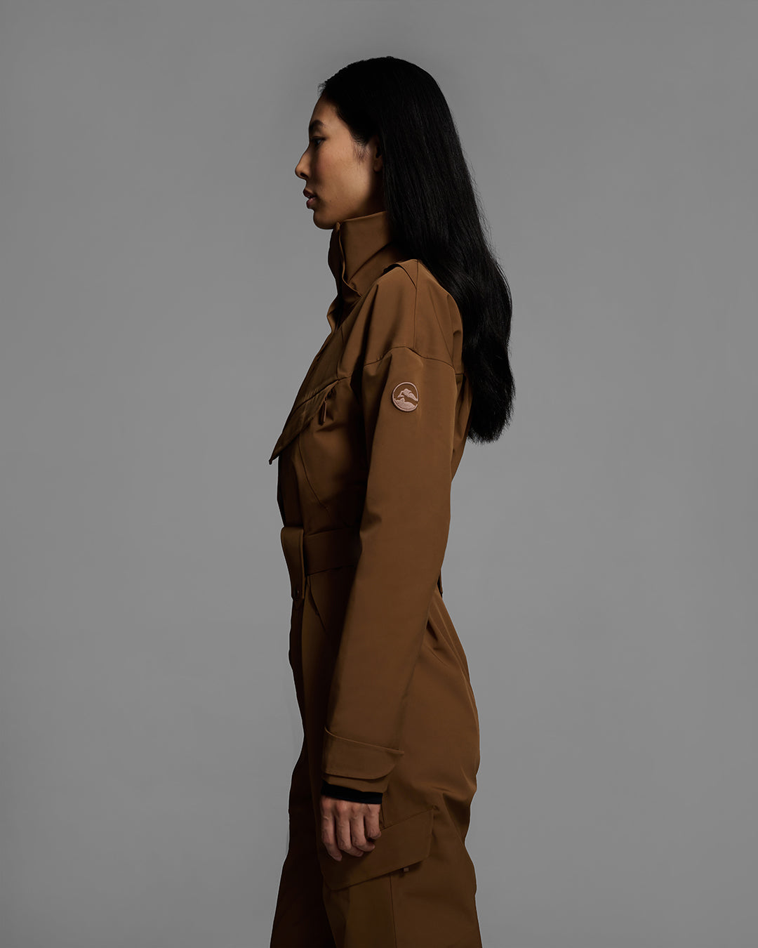 Side detail view of a female model wearing a belted sepia brown color ski suit made of technical waterproof fabric and zippers. #color_sepia