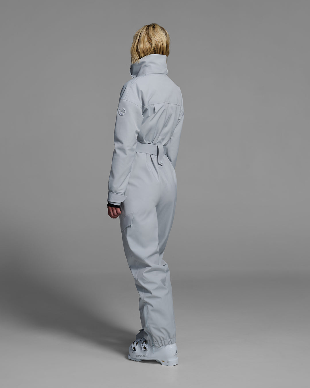 Back view of a female model wearing a belted smoke gray color ski suit made of technical waterproof fabric and zippers. #color_smoke