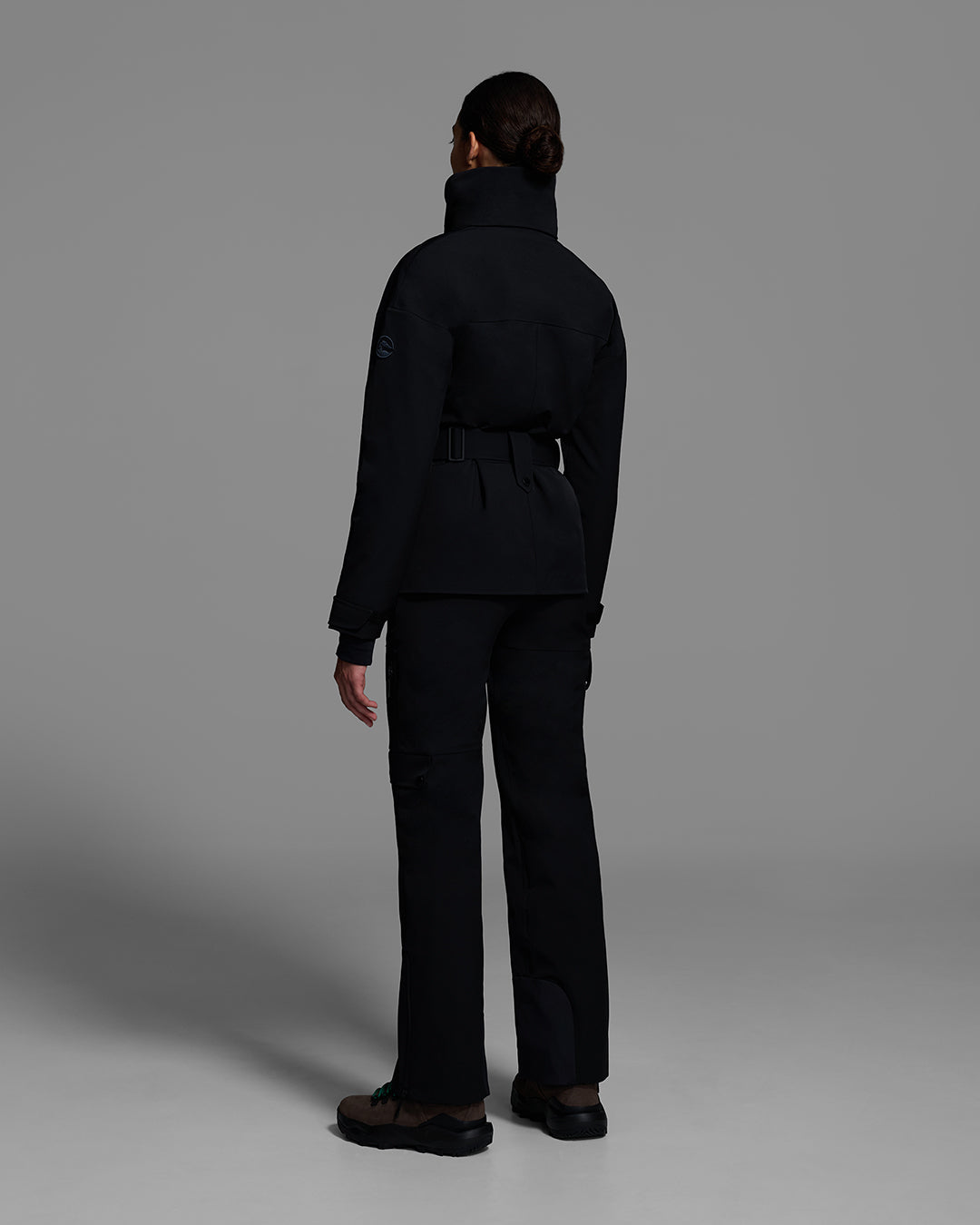 Back view of a female model wearing a belted ski jacket in onyx black made of technical waterproof fabric with waterproof zippers, and a matching black pant. #color_onyx