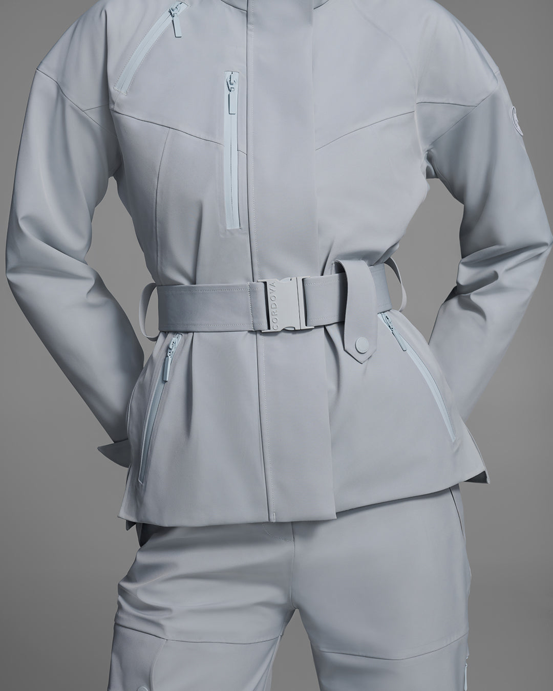 Detail view of a female model wearing a belted ski jacket in smoke gray technical waterproof fabric with waterproof zippers, and a matching gray pant. #color_smoke
