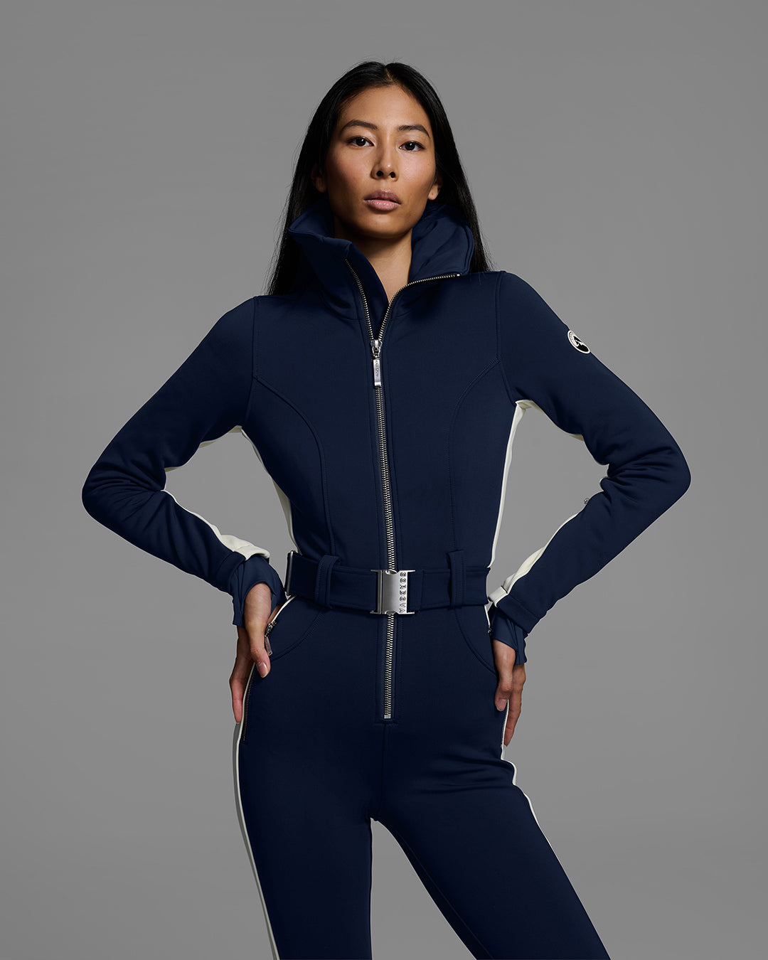 Front detail view of a female model wearing a body hugging ski suit in indigo blue with a white side stripe and belted waist. #color_indigo