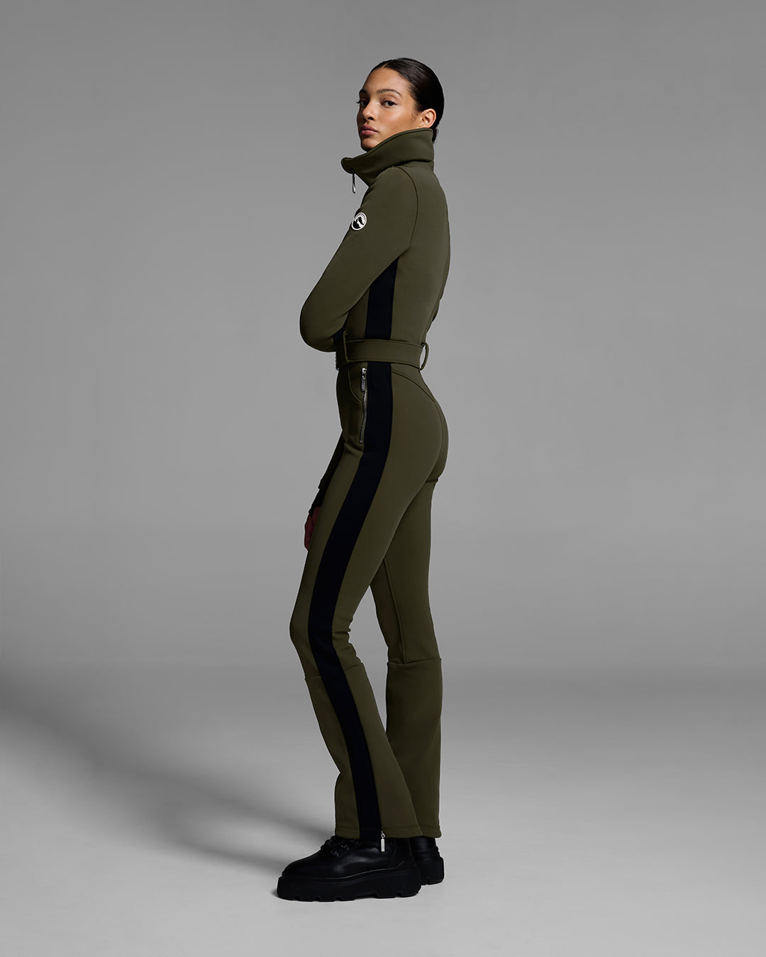 Side view of a female model wearing a body hugging ski suit in khaki green with a black side stripe and belted waist. #color_khaki