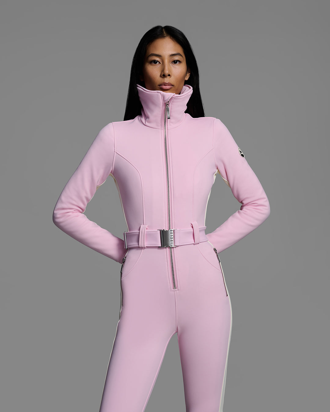 Detail front view of a female model wearing a body hugging ski suit in peony pink with a white side stripe and belted waist. #color_peony