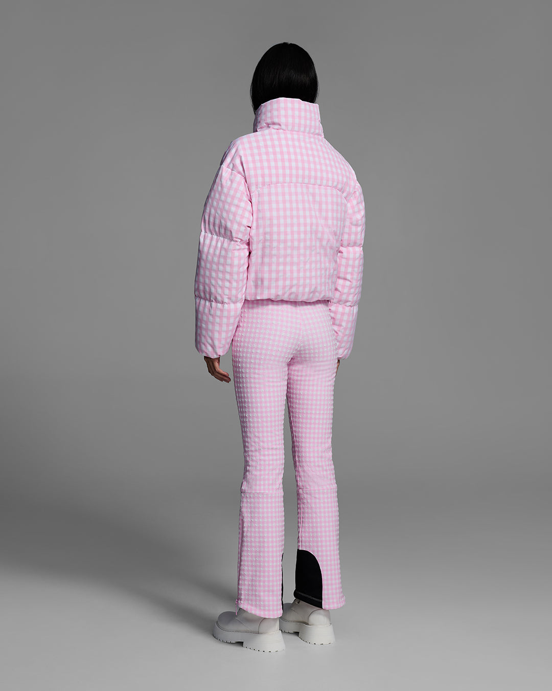 Back view of a female model wearing a down filled ski jacket in peony pink gingham fabric with matching ski suit underneath. #color_peony gingham