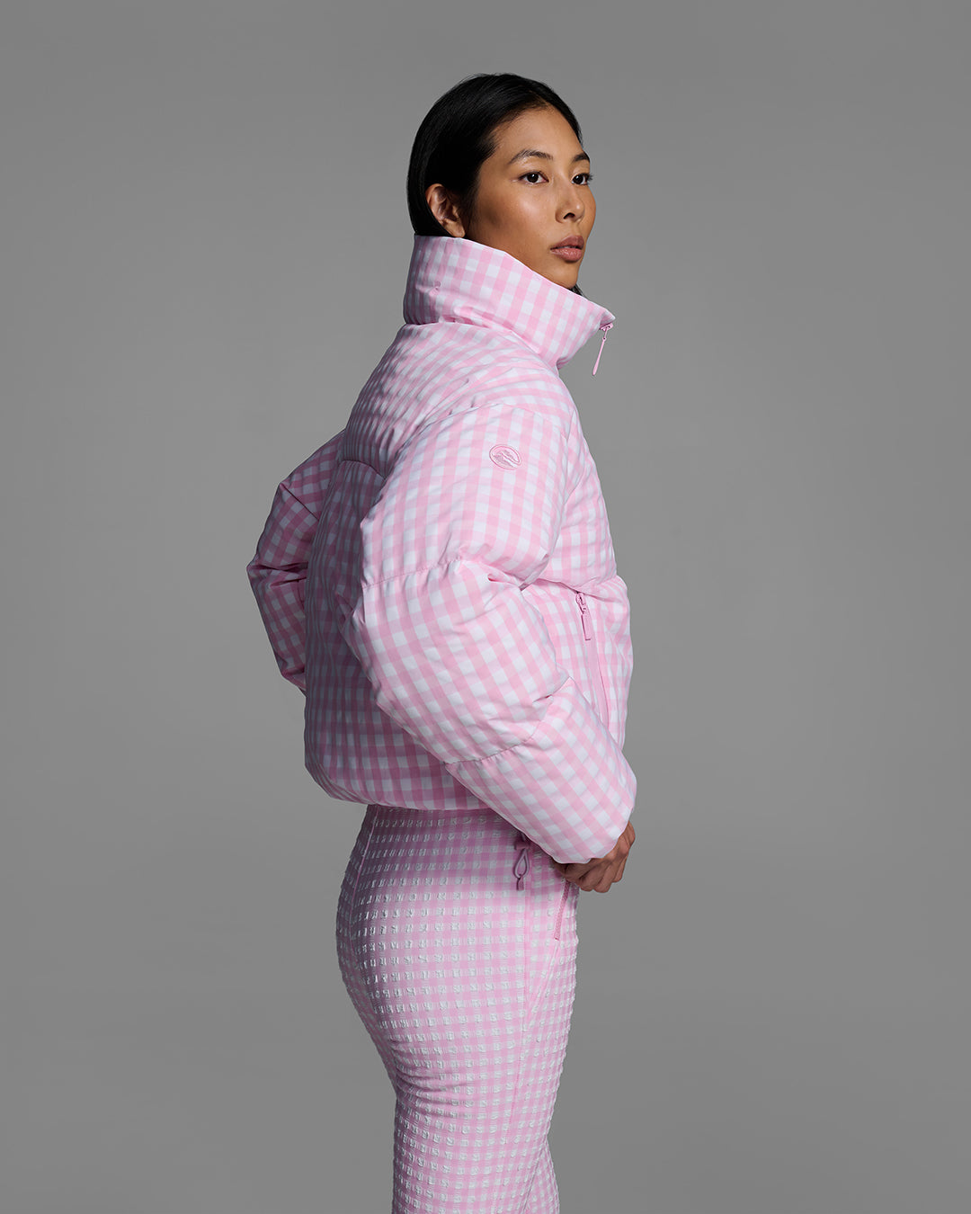 Side view of a female model wearing a down filled ski jacket in peony pink gingham fabric with matching ski suit underneath. #color_peony gingham