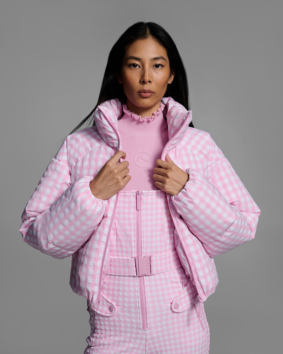 Front detail view of a female model wearing an unzipped down filled ski jacket in peony pink gingham fabric with matching ski bib underneath. #color_peony gingham