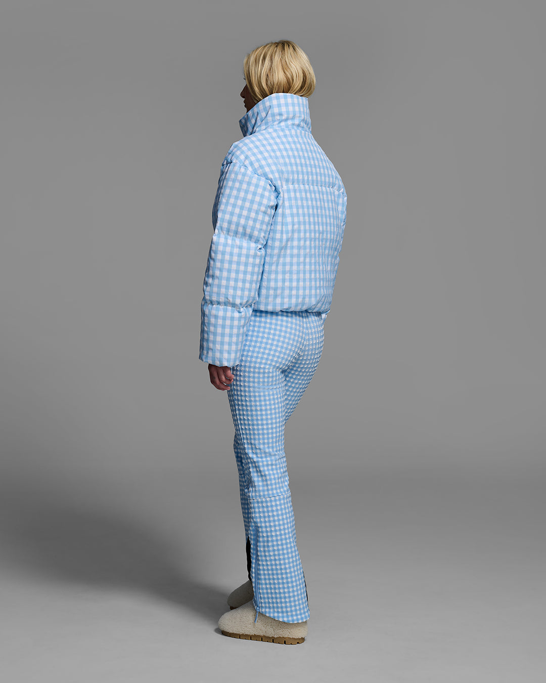 Back view of a female model wearing a down filled ski jacket in cerulean blue gingham fabric with matching ski suit underneath. #color_cerulean gingham