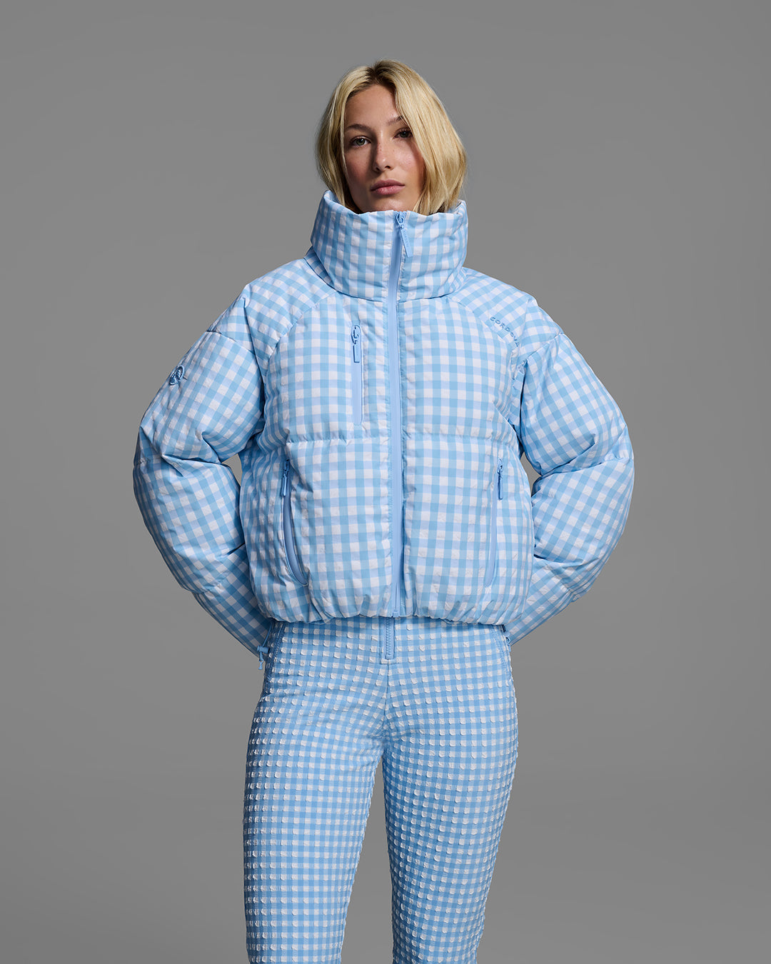 Front detail of a female model wearing a down filled ski jacket in cerulean blue gingham fabric with matching ski suit underneath. #color_cerulean gingham