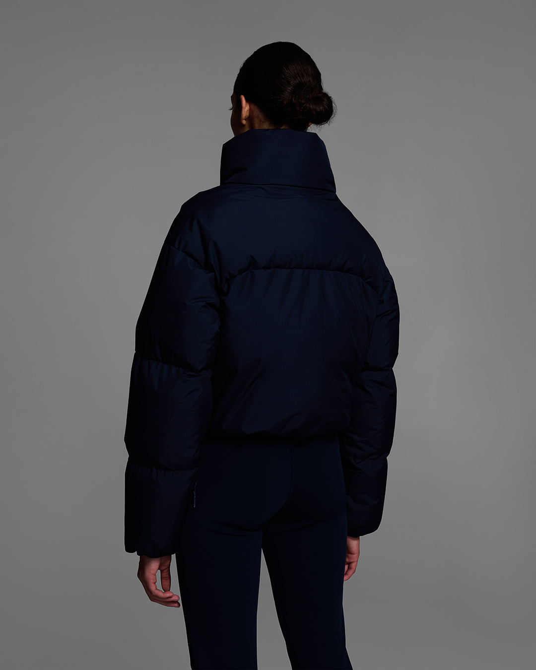 Back view of a female model wearing a down filled ski jacket in indigo blue fabric with a matching ski bib underneath. #color_indigo