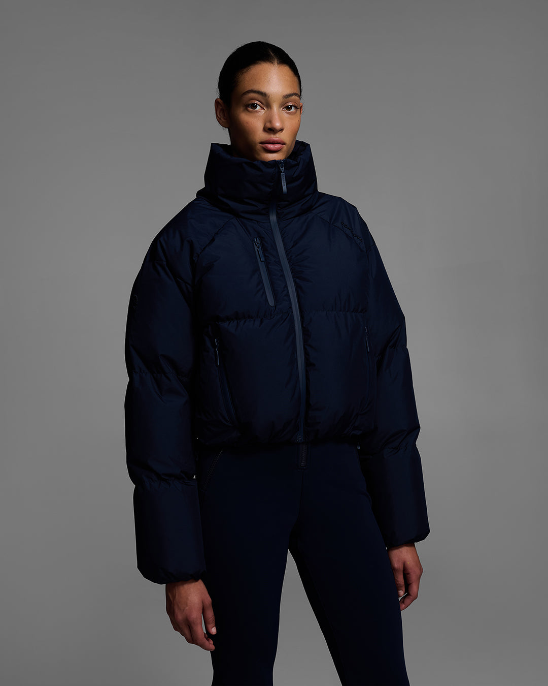 Female model wearing a down filled ski jacket in indigo blue fabric zipped up with a matching ski bib underneath. #color_indigo