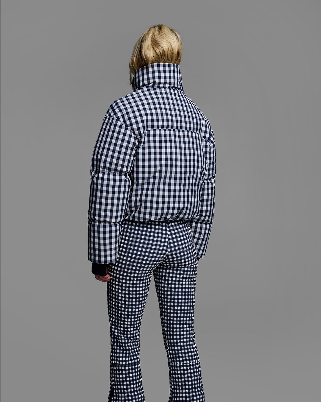 Back view of a female model wearing a down filled ski jacket in indigo blue gingham fabric with a matching ski bib underneath. #color_indigo gingham