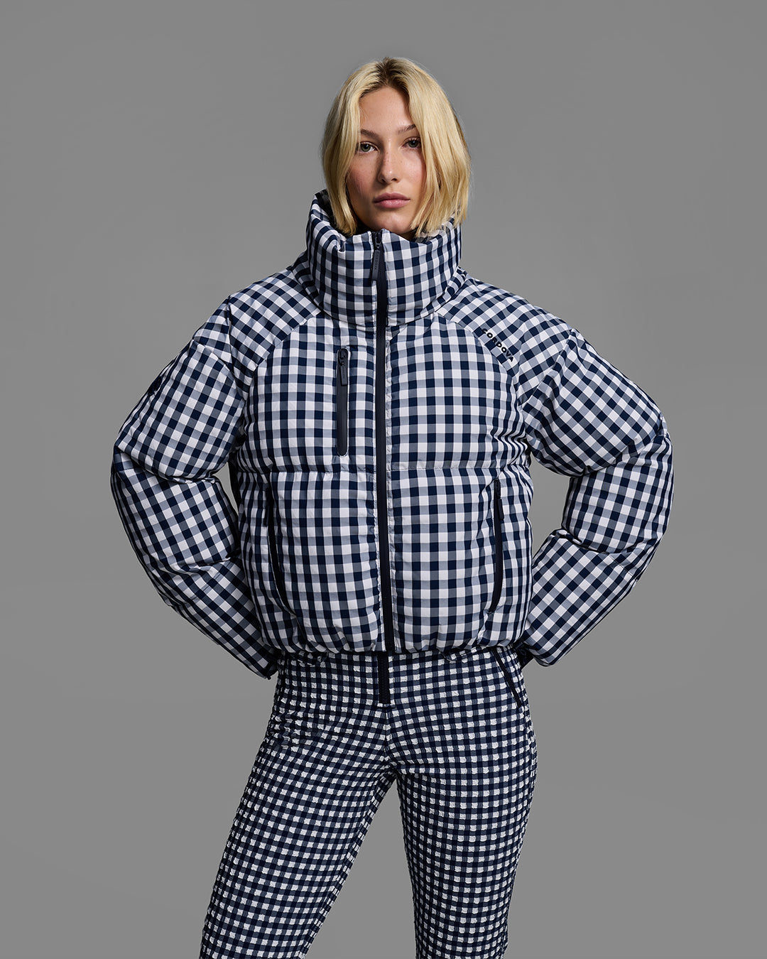 Front detail view of a female model wearing a down filled ski jacket in indigo blue gingham fabric with a matching ski bib underneath. #color_indigo gingham