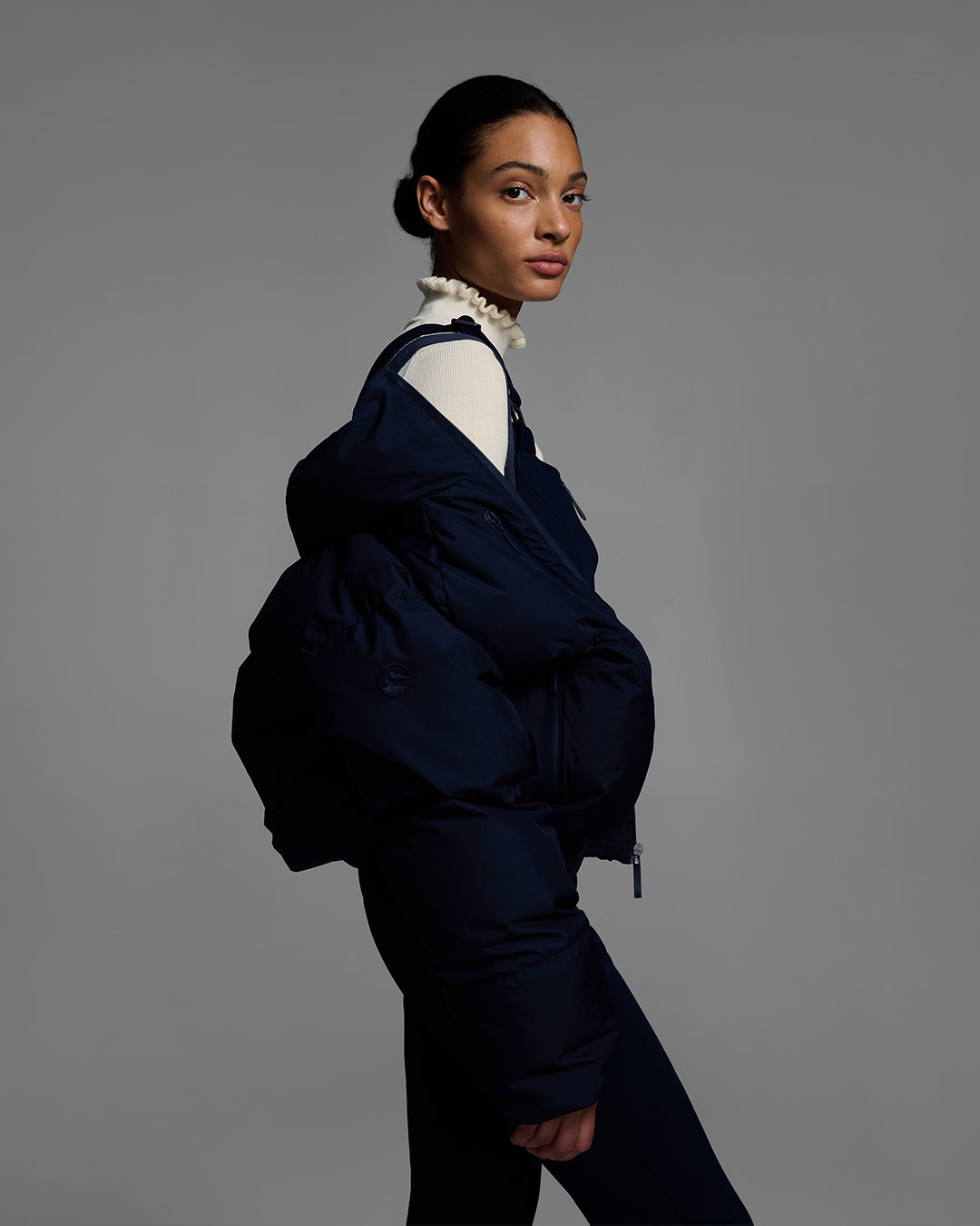 Female model wearing a down filled ski jacket in an indigo blue fabric, hanging off the shoulders to highlight the backpack straps feature. #color_indigo