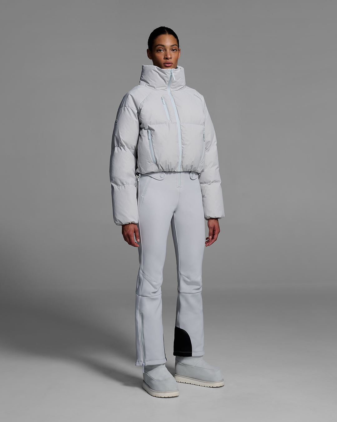 Female model wearing a down filled ski jacket in smoke gray fabric, zipped up with a matching ski bib underneath. #color_smoke