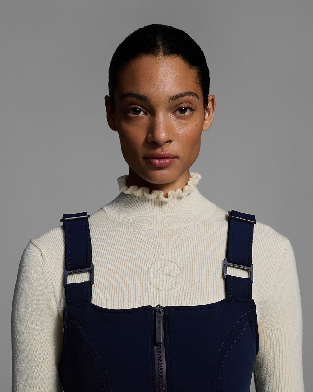 Close up of a female model wearing a belted indigo blue color ski bib with complementary white sweater, highlighting the bib straps. #color_indigo
