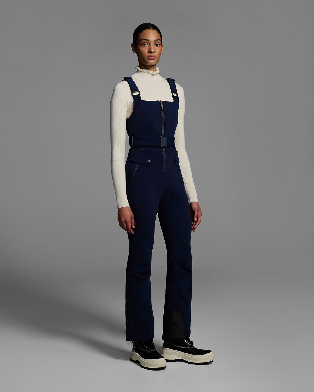 Female model wearing a belted indigo blue color ski bib with complementary white sweater. #color_indigo