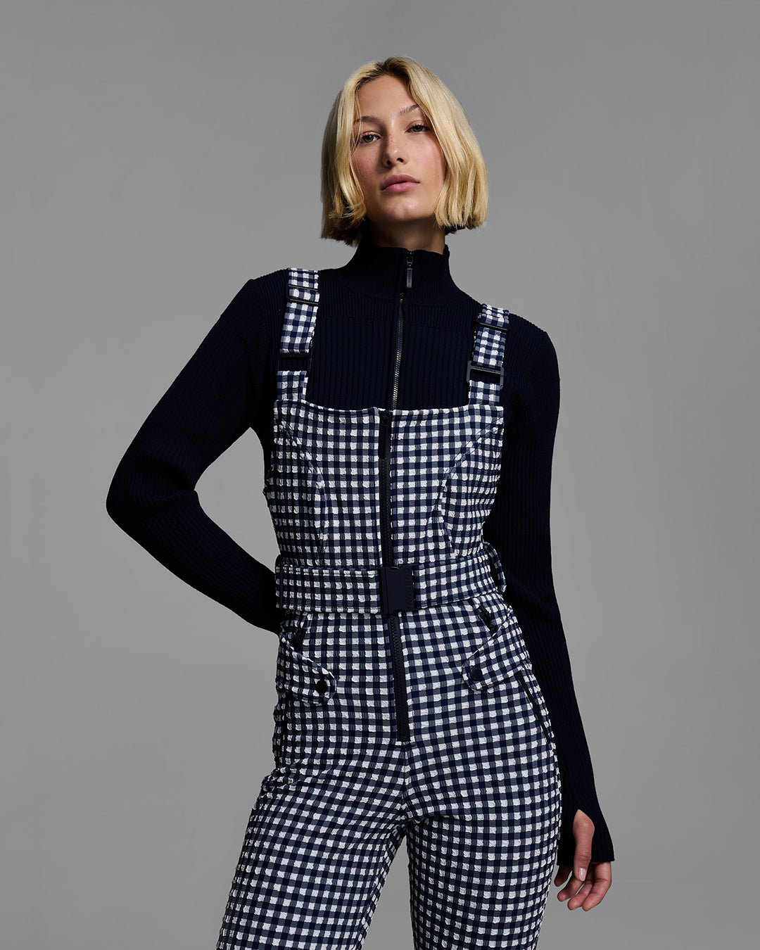 Torso detail shot of a female model wearing a belted indigo blue color ski bib in a gingham print with dark blue sweater. #color_indigo gingham