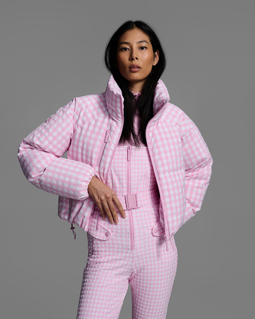 Female model wearing a belted peony pink color ski bib in a gingham print with a matching ski jacket over the top, unzipped. #color_peony gingham