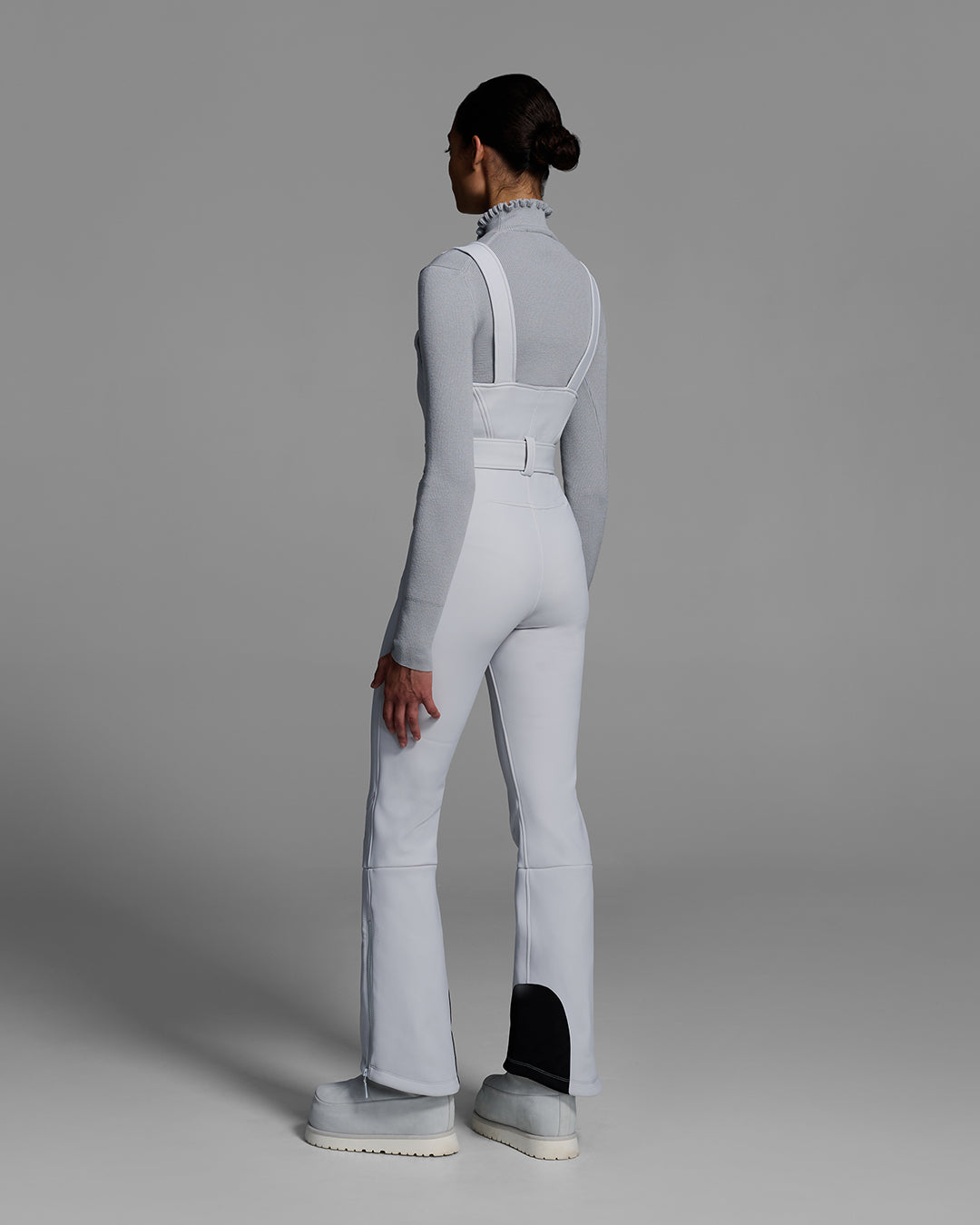 Back view of a female model wearing a belted smoke gray color ski bib with a matching gray sweater underneath. #color_smoke