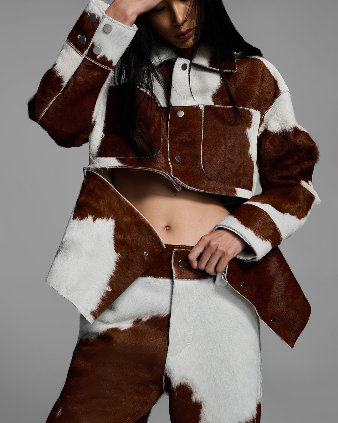 Detail view of a female model wearing a matching jacket and pant set crafted in hair-on cowhide leather with the jacket unzipped showing a panel can be removed to be a crop jacket. #color_cowhide