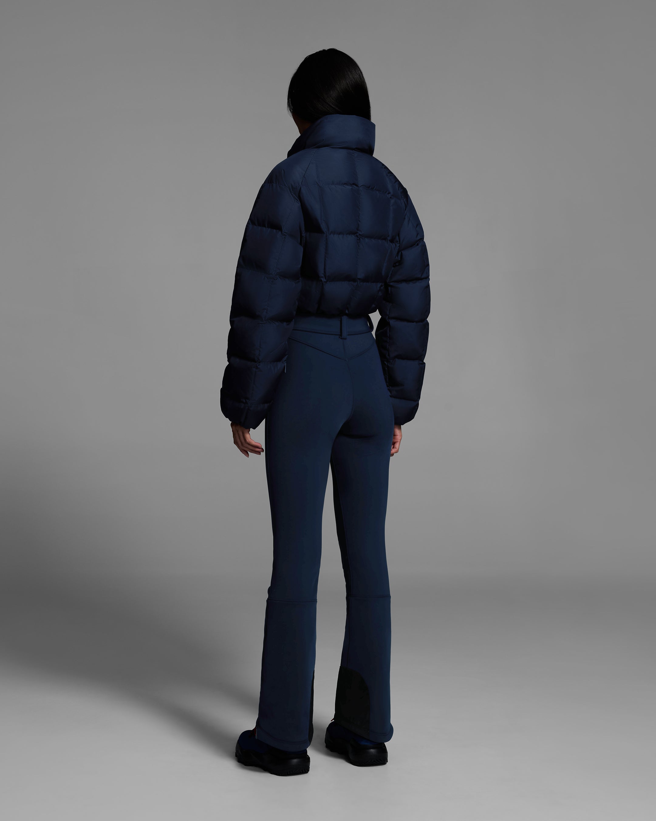 Back view of a female model wearing a belted indigo blue color ski suit with quilted, down filled top and 4-way stretch bottom. #color_indigo