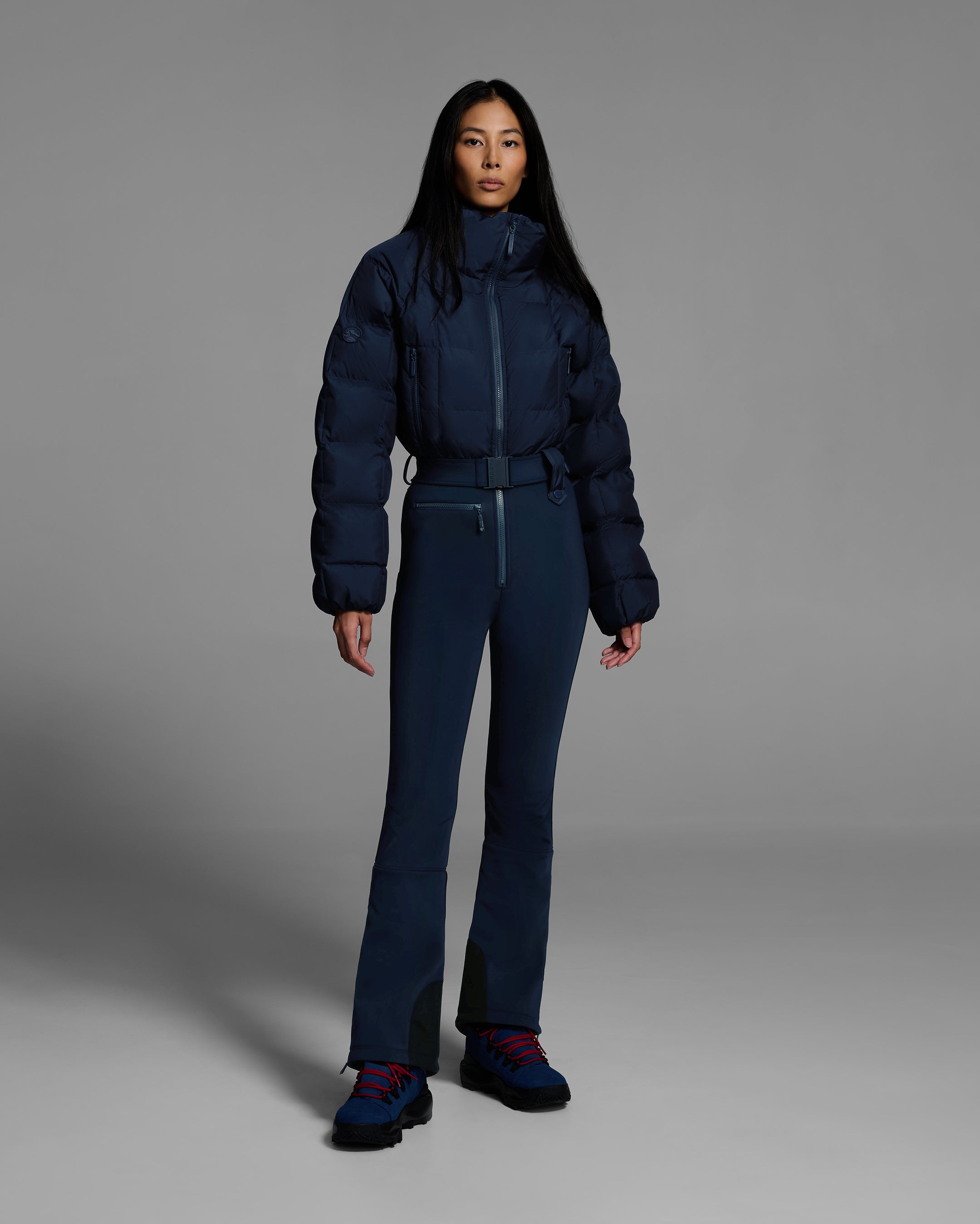 Female model wearing a belted indigo blue color ski suit with quilted, down filled top and 4-way stretch bottom. #color_indigo