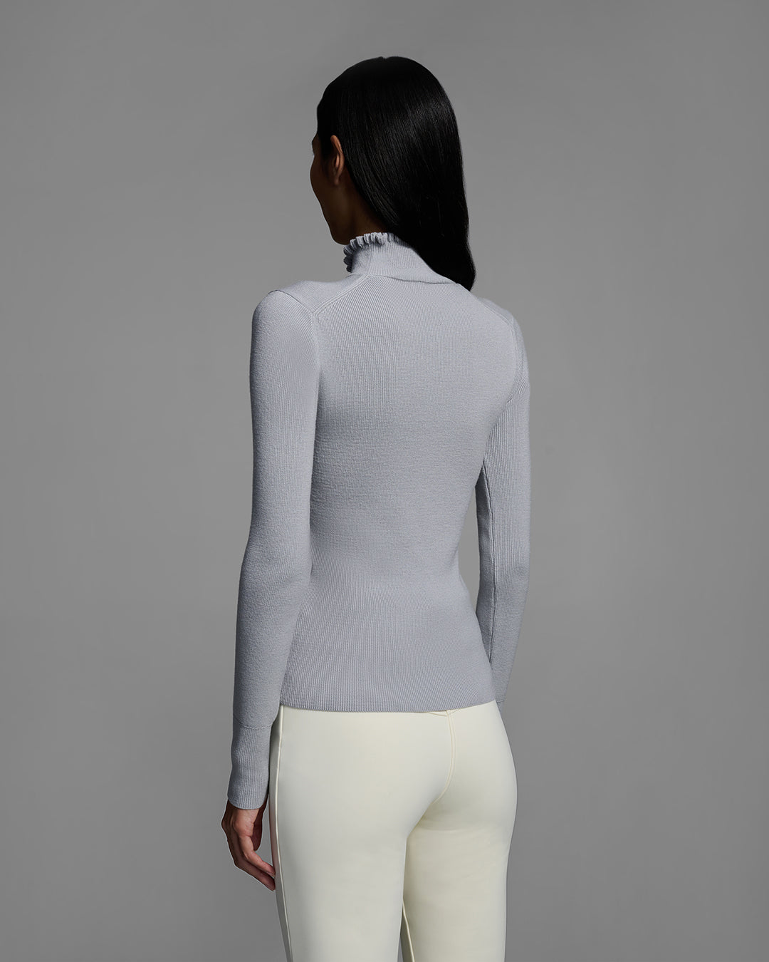 Back view of a female model wearing a gray melange knit ski sweater with ruffle edged turtleneck and 3d mountain logo on chest with white ski pant. #color_gray melange