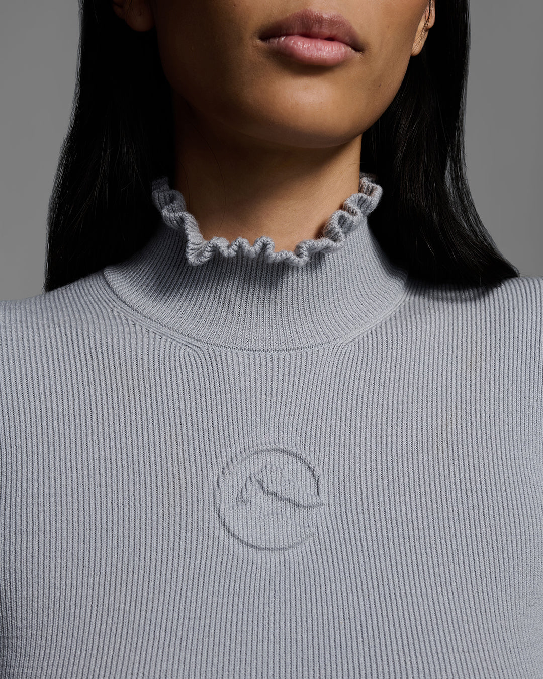 Detail shot of a Female model wearing a gray melange knit ski sweater with ruffle edged turtleneck and 3d mountain logo on chest. #color_gray melange