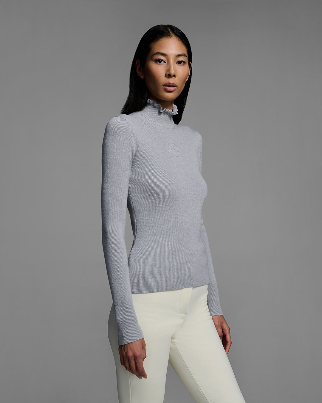 Female model wearing a gray melange knit ski sweater with ruffle edged turtleneck and 3d mountain logo on chest with white ski pant. #color_gray melange