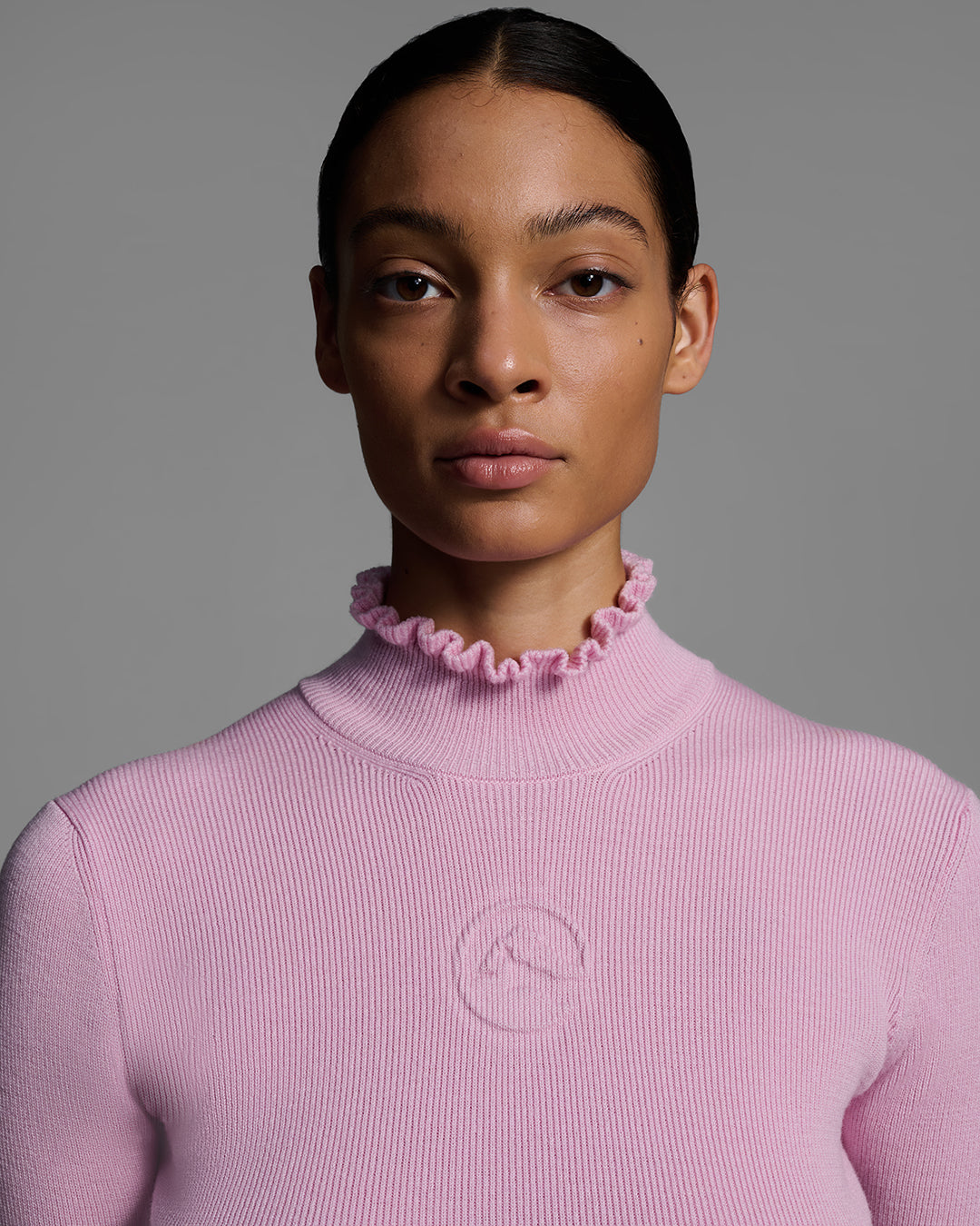 Close up detail view of a female model wearing a peony pink knit ski sweater with ruffle edged turtleneck and 3d mountain logo on chest. #color_peony
