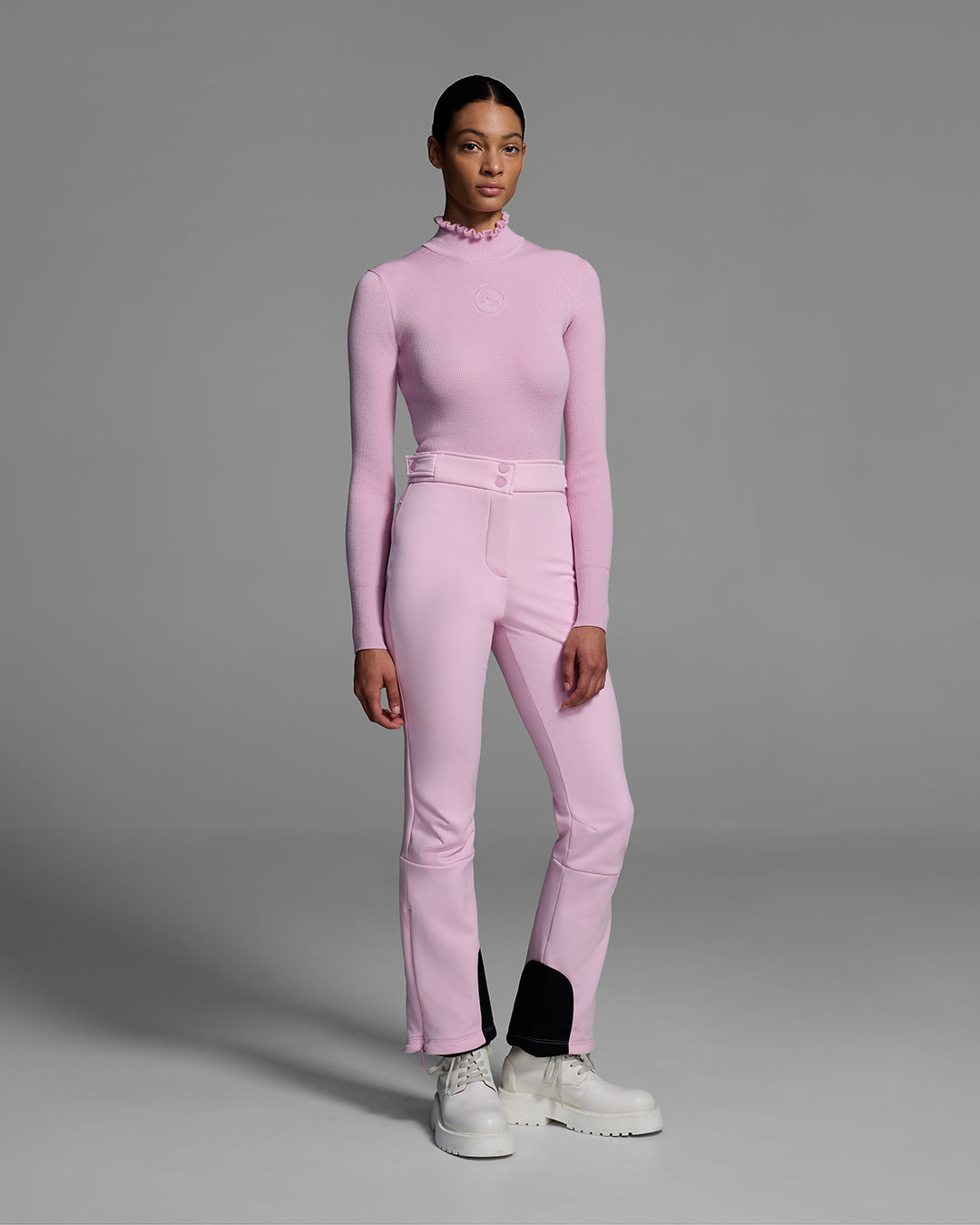 Female model wearing a peony pink knit ski sweater with ruffle edged turtleneck and 3d mountain logo on chest with matching pink ski pant. #color_peony