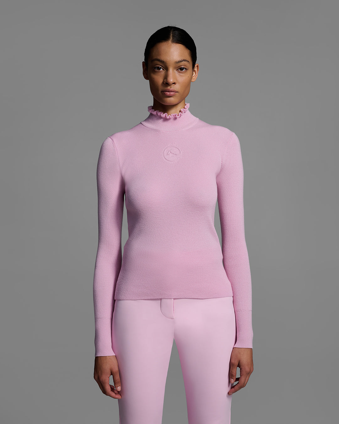 Female model wearing a peony pink knit ski sweater with ruffle edged turtleneck and 3d mountain logo on chest with matching pink ski pant. #color_peony