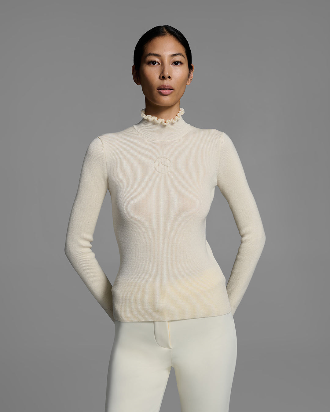 Woman wearing a white ski sweater with a high ruffle neck and 3D mountain logo on chest. #color_alabaster