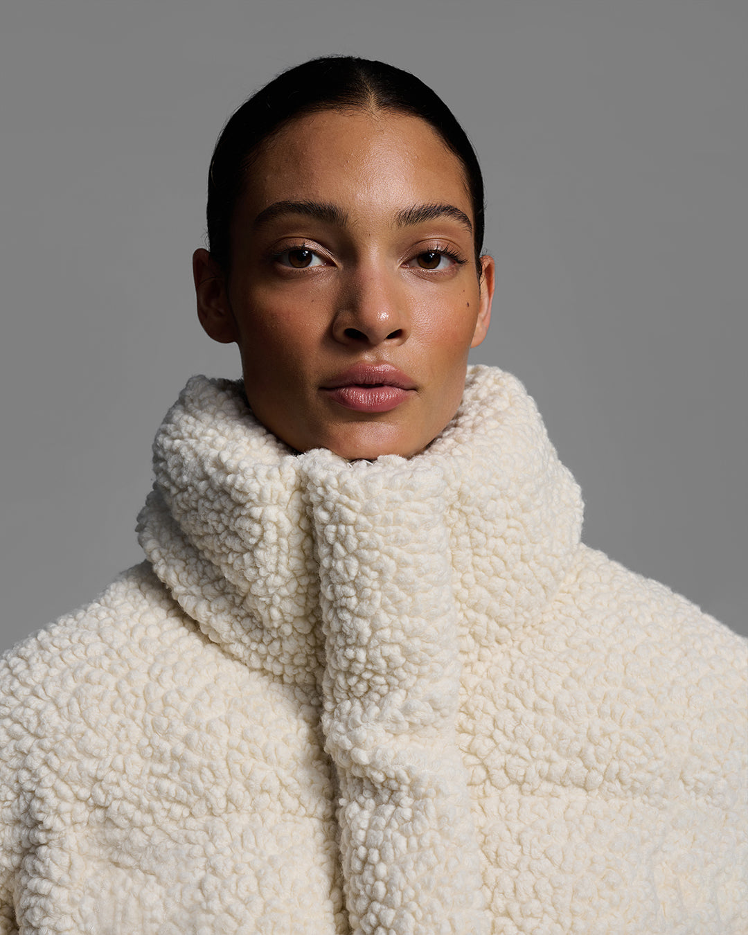 Detail view of female model wearing a natural, off-white, sherpa puffer on a gray background. #color_natural
