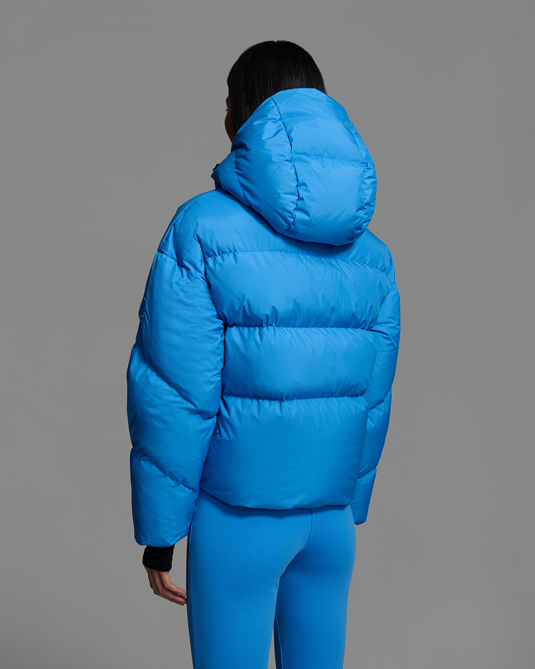 Back view of a female model wearing a classic down filled ski jacket in bright blue with a matching ski pant. #color_azul