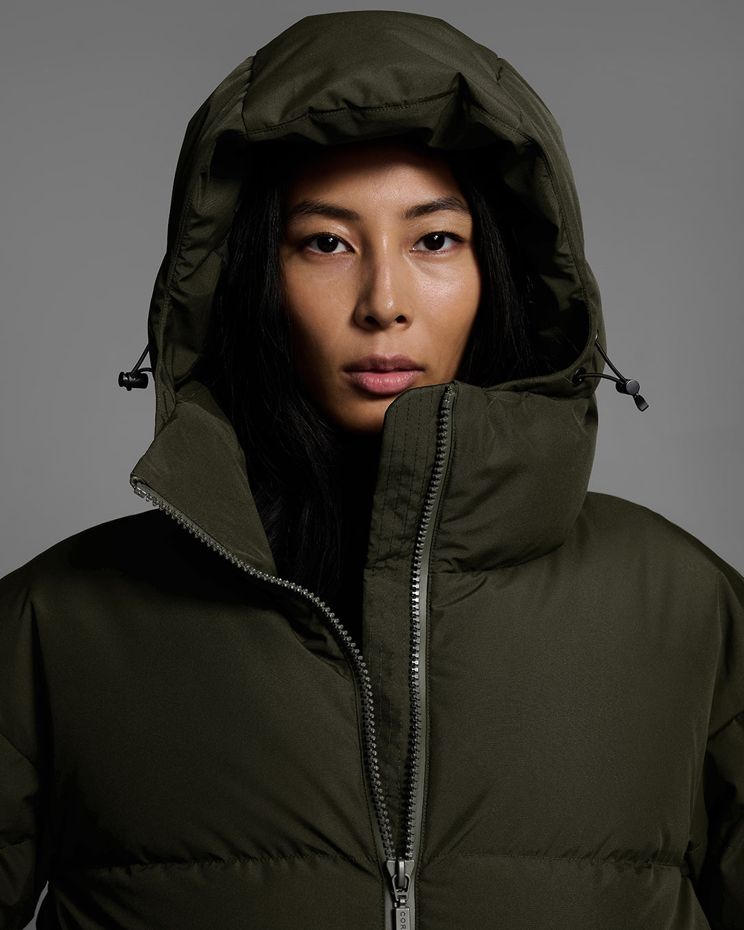 Close up detai of the hood of a female model wearing a classic down filled ski jacket in khaki green. #color_khaki