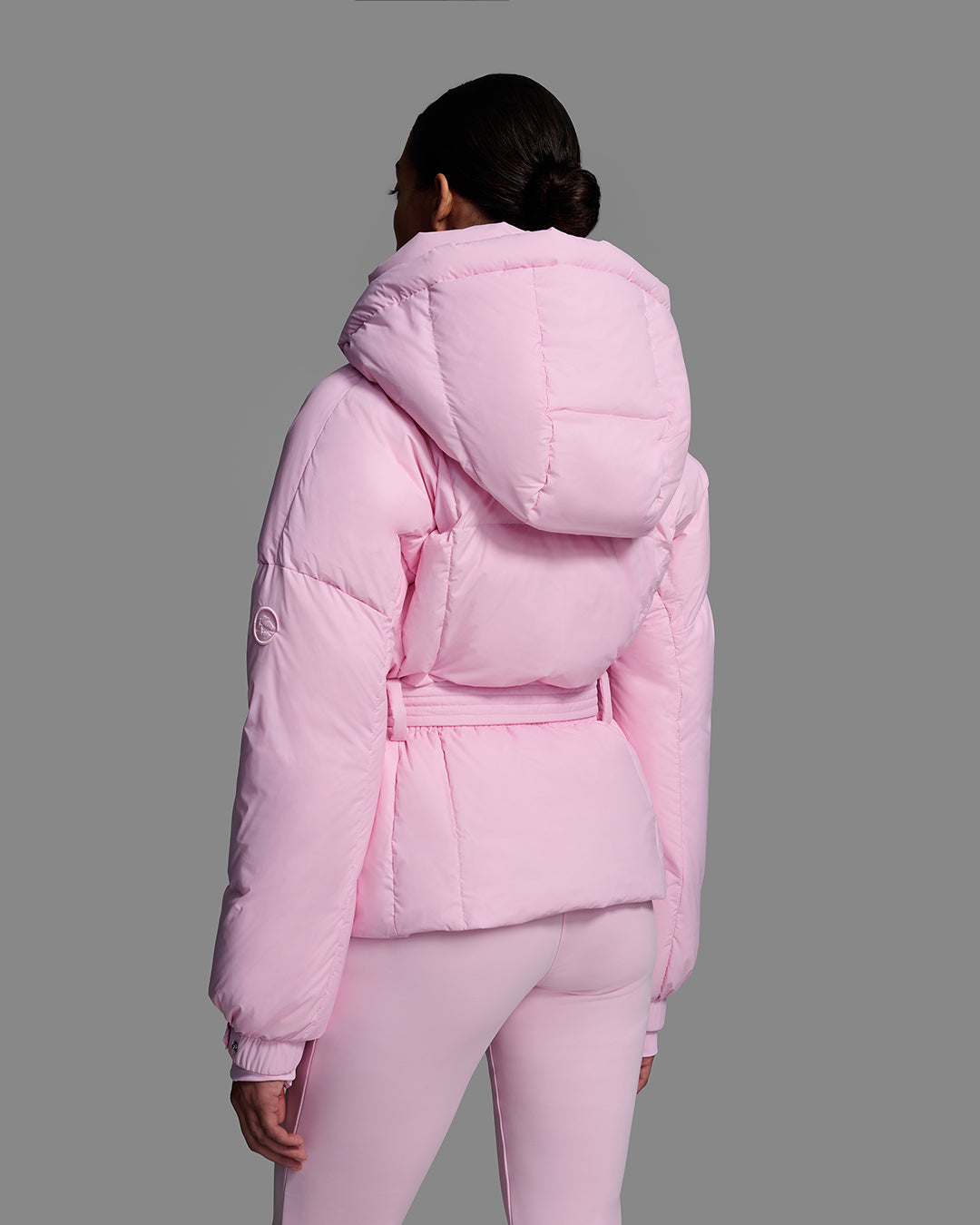 Back view of a female model wearing a classic slope style down filled, belted ski jacket in peony pink color with a matching ski pant. #color_peony