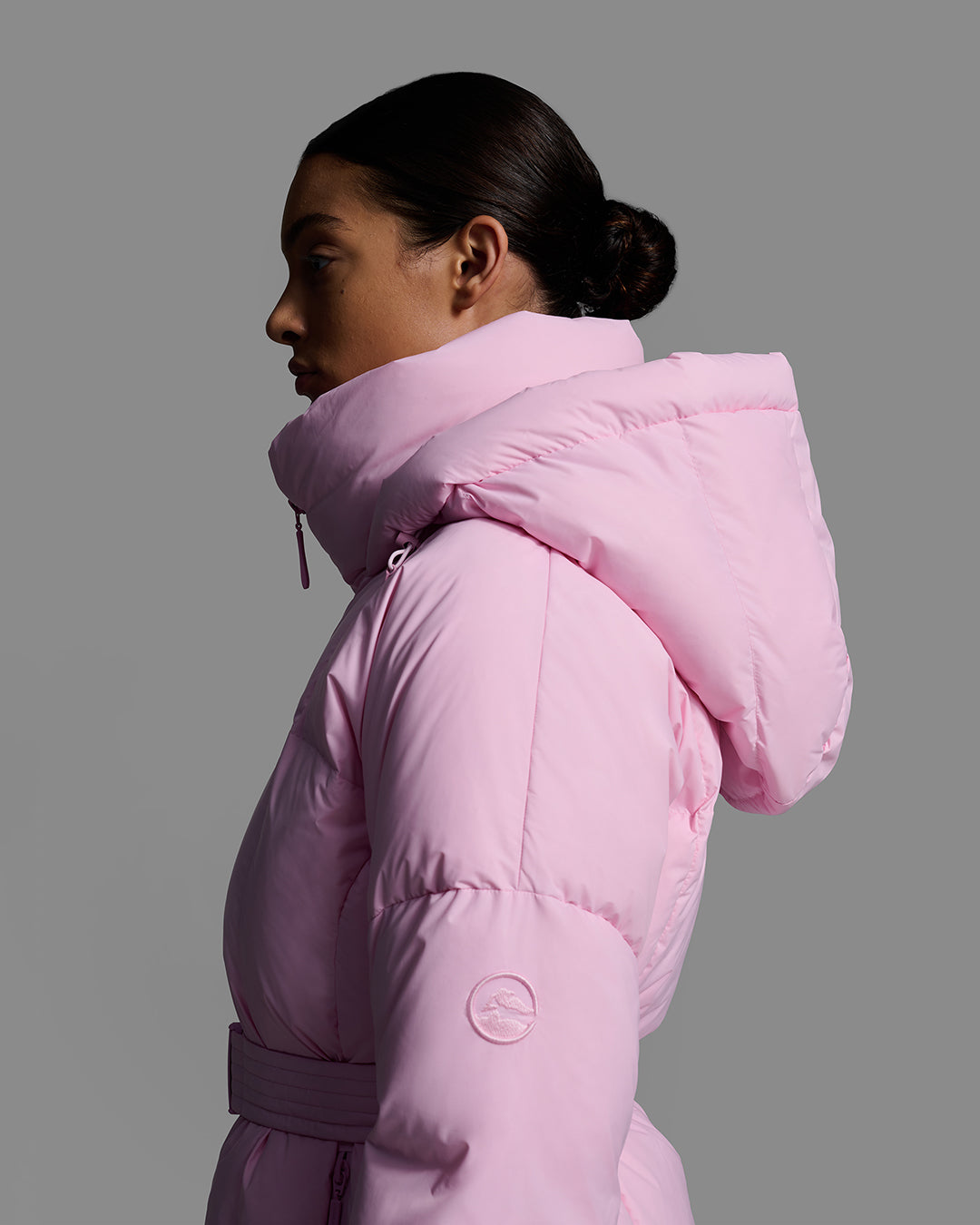 Detail side view of a female model wearing a classic slope style down filled, belted ski jacket in peony pink color. #color_peony