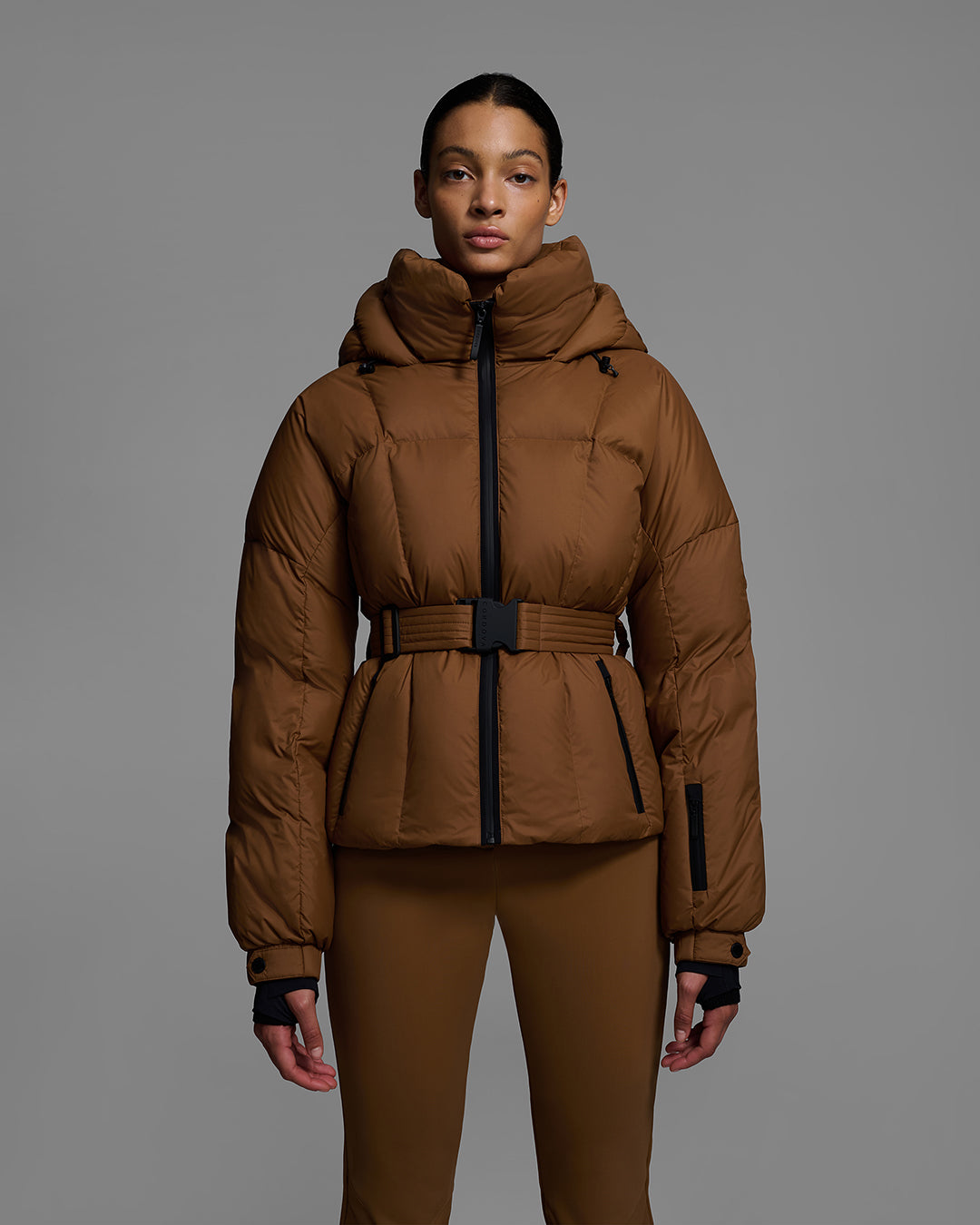Detail front view of a female model wearing a classic slope style down filled, belted ski jacket in sepia brown color with a matching ski pant. #color_sepia