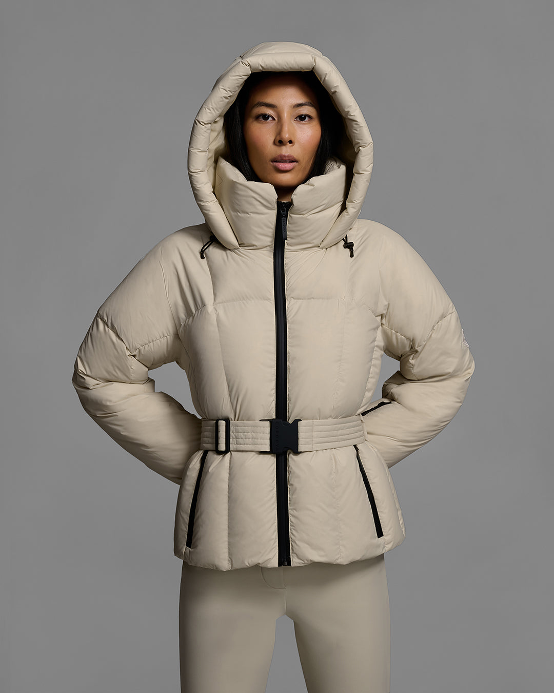 Detail front view of a female model wearing a classic slope style down filled, belted ski jacket with the hood up, in birch inspired tan color with a matching ski pant. #color_birch