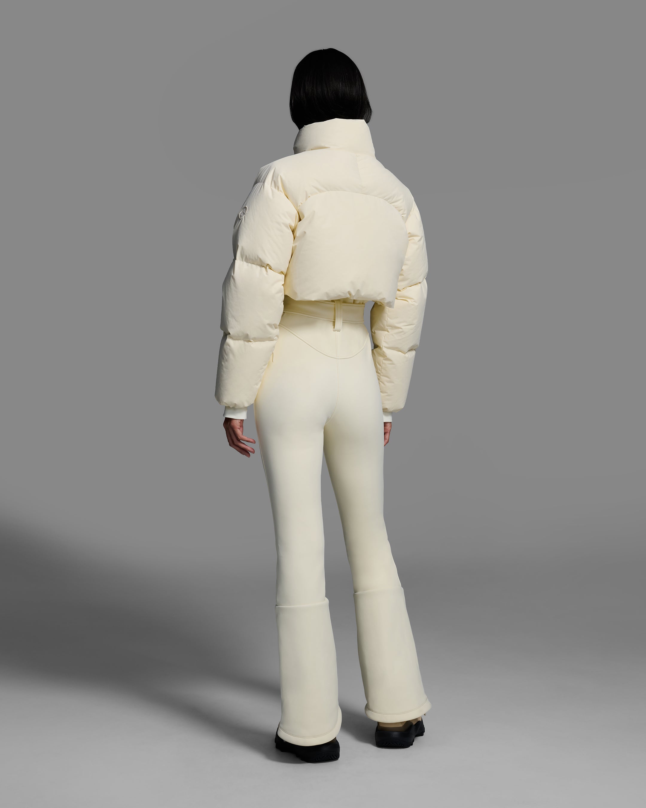 Back view of a female model wearing an alabaster white ski suit with a white puffer crop top, belted waist, and 4-way stretch pant. #color_bone