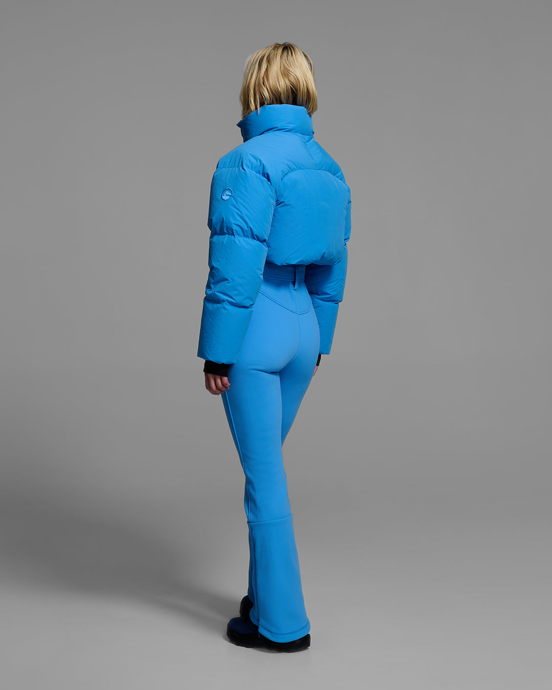 Back view of a female model wearing an bright blue ski suit with a puffer crop top, belted waist, and 4-way stretch pant. #color_azul