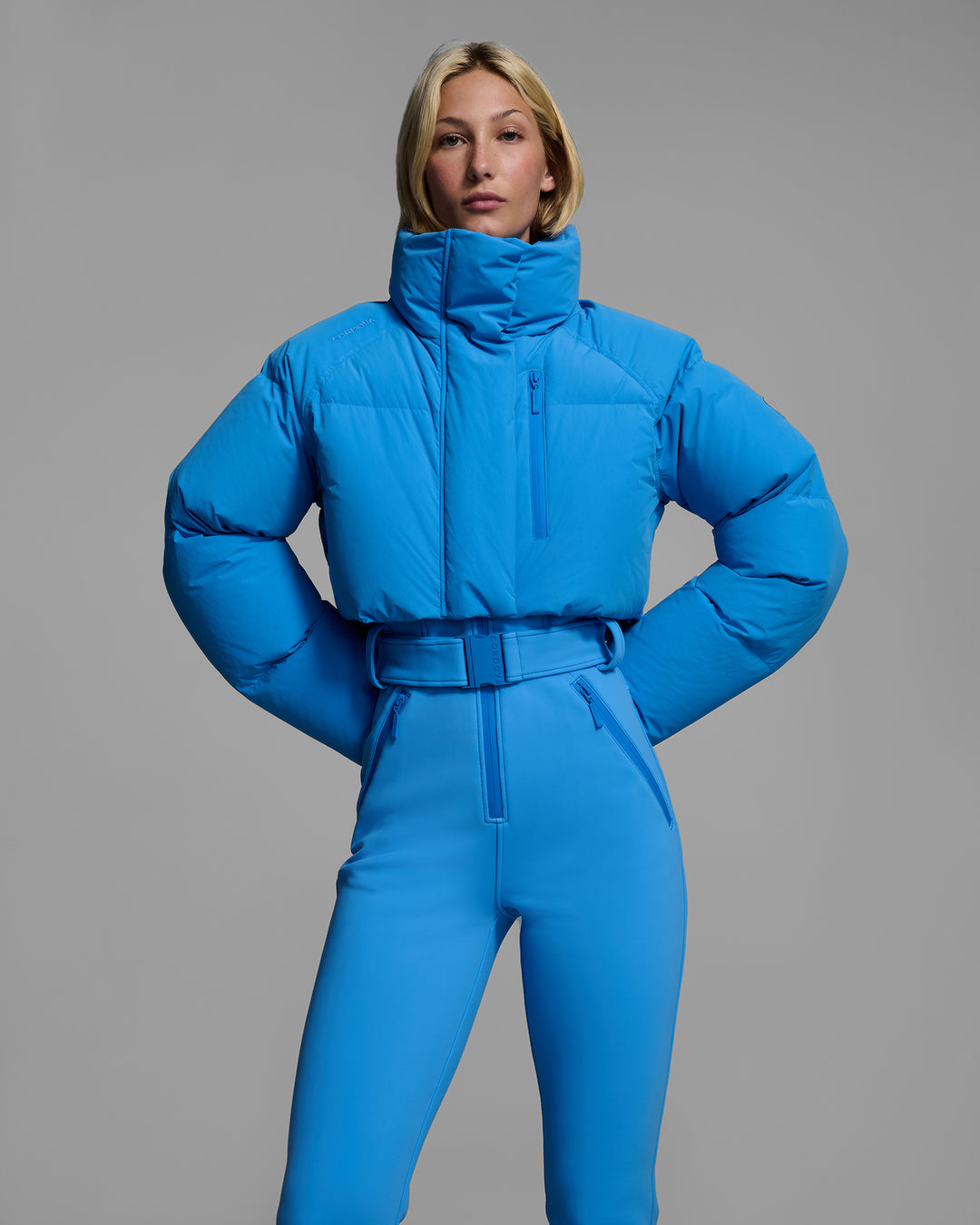 Detail front view of a female model wearing an bright blue ski suit with a puffer crop top, belted waist, and 4-way stretch pant. #color_azul