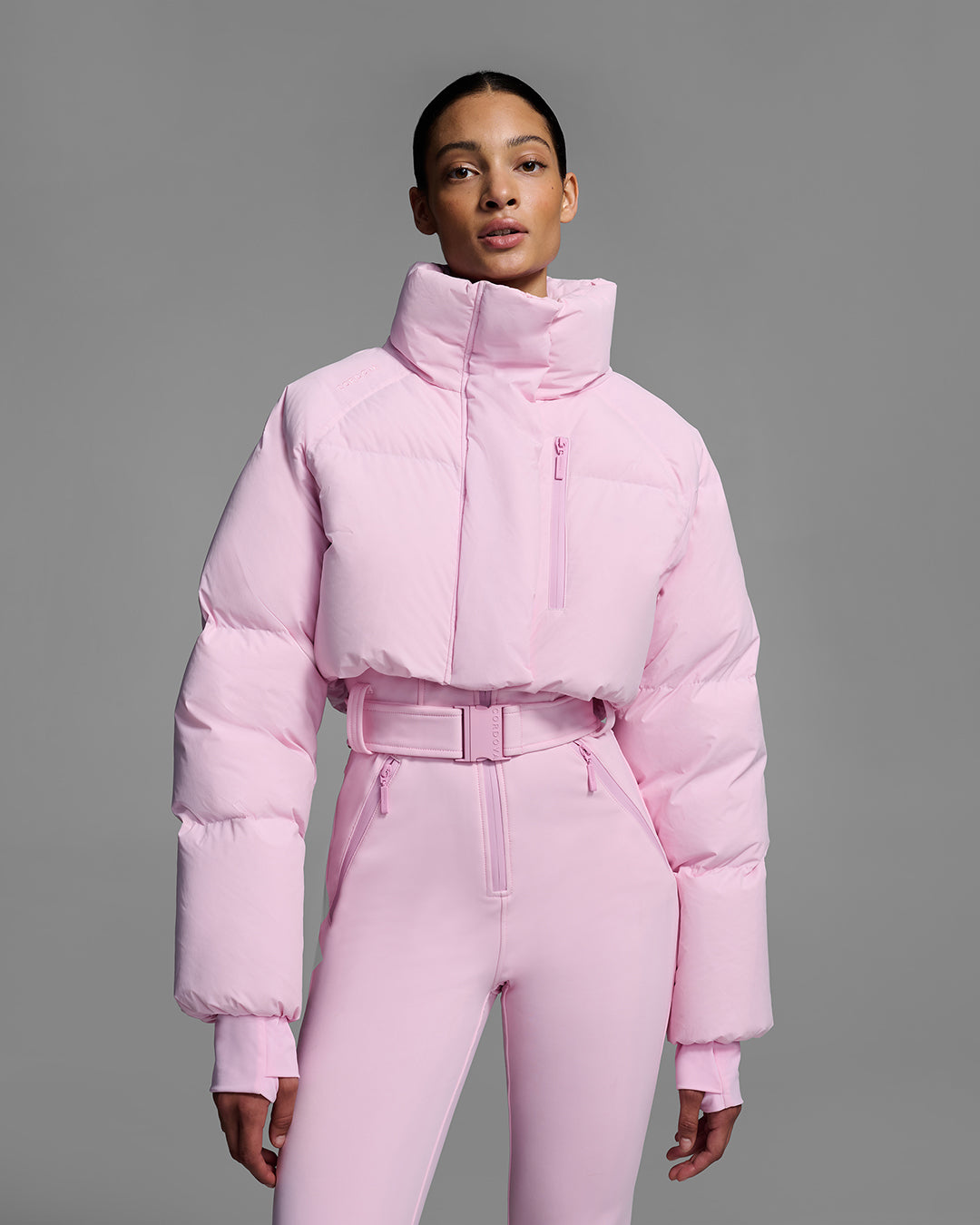 Detail view of a female model wearing a peony pink ski suit with a puffer crop top, belted waist, and 4-way stretch pant. #color_peony